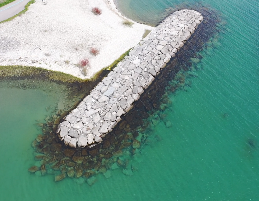 An aerial view of a short breakwater-like structure called a headland. It is made of large flat stones and is in good condition. The headland is connected to the shoreline by cobbles, and a trail can be seen at the upper left. The water surrounding the headland is transparent and the underwater section of the structure is clearly visible.