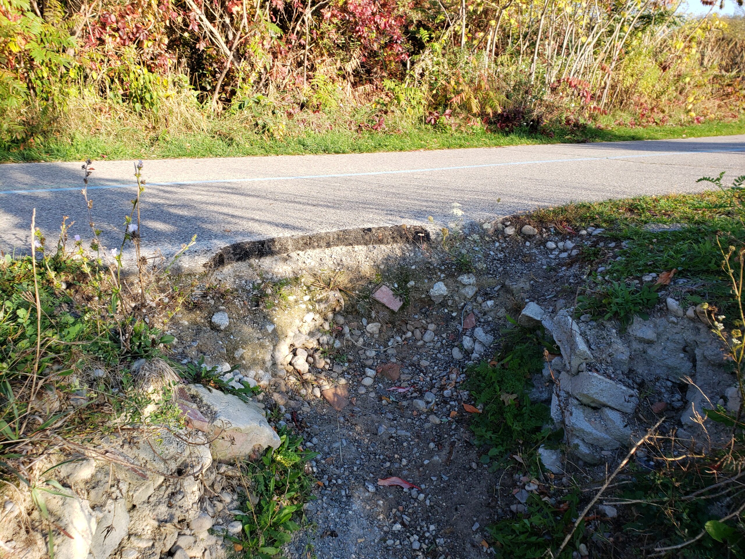 A close-up of a portion of the paved trail, showing a broken section along the edge. The slope immediately below the broken portion is exposed soil with no vegetation due to erosion.