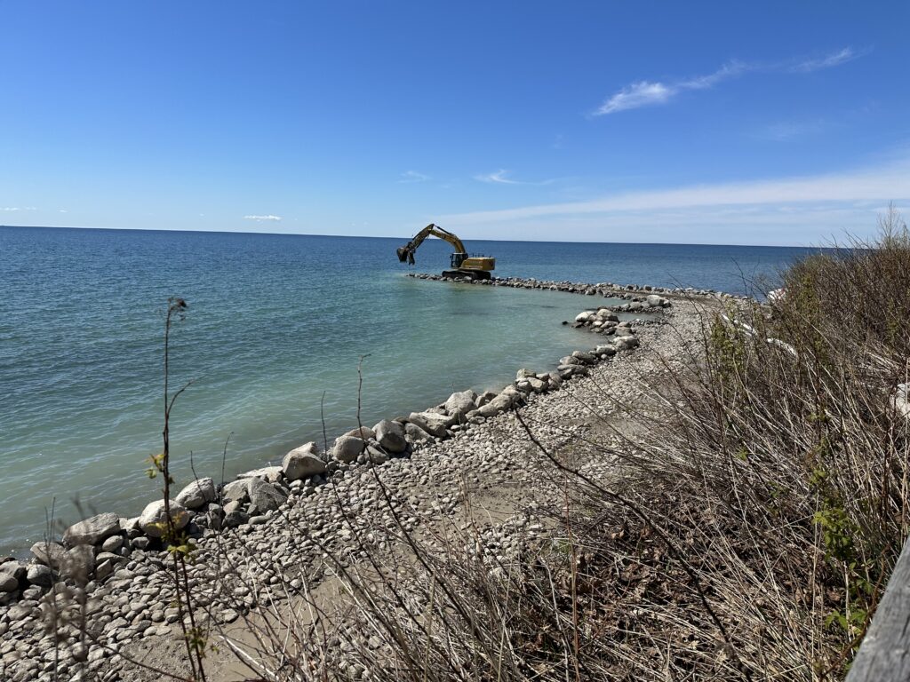 An access road follows the shoreline before extending into Lake Ontario. An excavator is stationed at the end of the road, placing stone material into the lake.