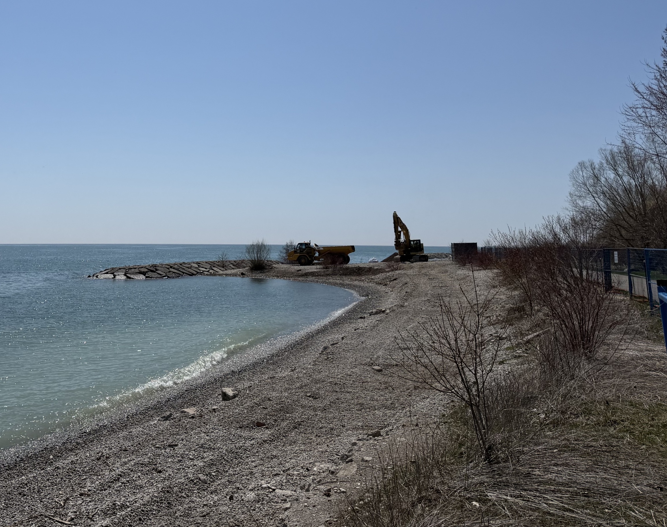 An excavator and truck are visible near a headland along the shoreline. Lake Ontario is visible on the left, and the cobble shoreline is in the foreground on the right.