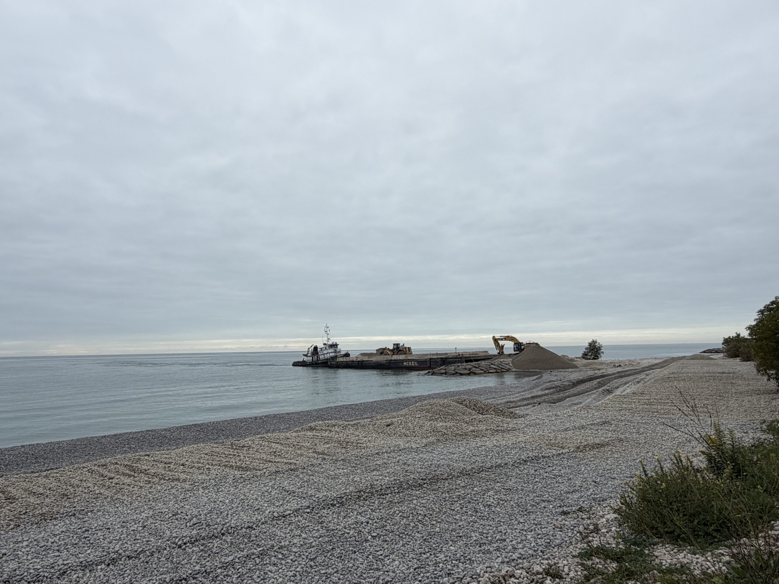 A barge carrying cobble is docked at the headland in the background, with an excavator working to place the cobble on the shoreline. The shoreline is visible in the foreground partially completed. Mounds and ruts are visible in the cobble profile.